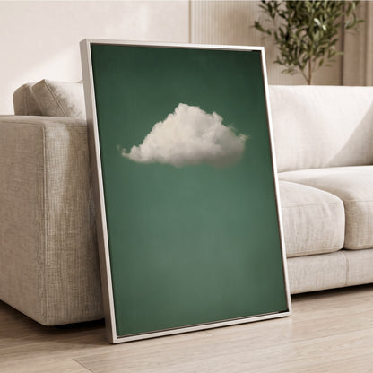 A framed wall art print leaning against a light beige sofa in a living room. The print shows a single white cloud centered on a muted green background, with a thin metallic frame.