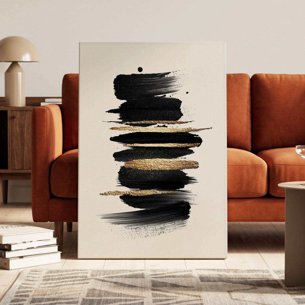 Black Gold Brushstrokes: abstract canvas wall art with black and textured gold brushstrokes on a cream background, displayed leaning against an orange-brown sofa in a modern living room setting.