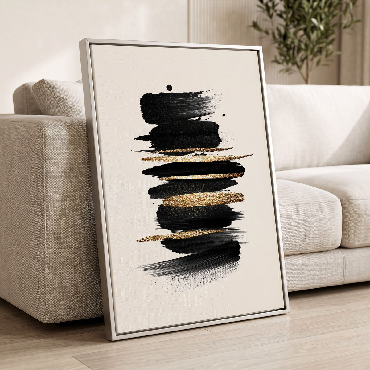 Black Gold Brushstrokes: a framed abstract wall art print leaning against a light beige sofa in a modern living room. The artwork features bold horizontal black brushstrokes with textured gold metallic accents on an off-white background, enclosed in a thin silver frame.