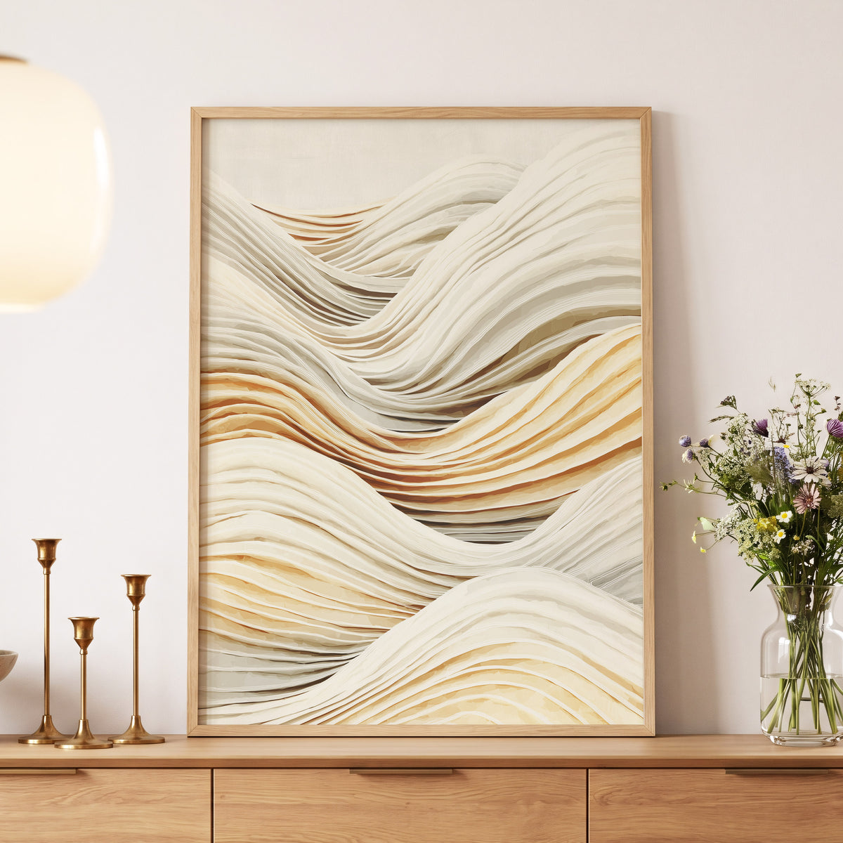 Ivory Layered Waves: a framed abstract wall art print with layered wavy lines in cream, beige, and warm tan tones, displayed on a wooden console against a light wall with decorative candles and a flower vase nearby.
