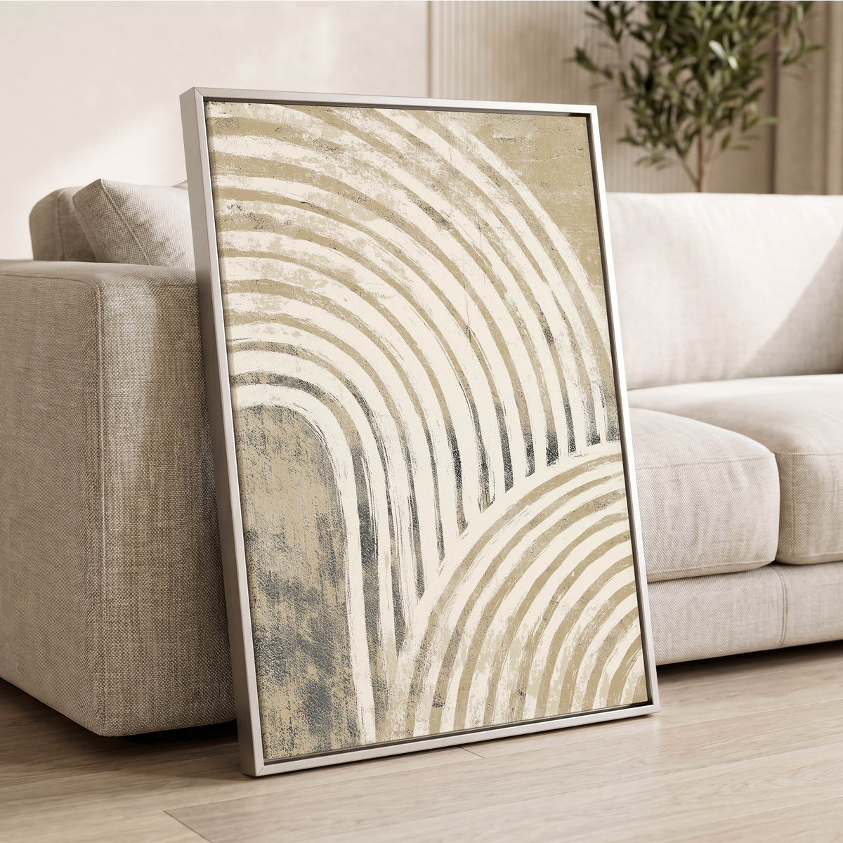 Neutral Textured Arches: a framed abstract wall art print leaning against a light beige sofa in a living room setting. The artwork features flowing, curved lines in cream, beige, and muted gray tones, enclosed in a thin silver frame.