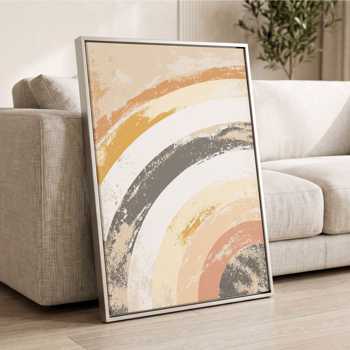 Pastel Rainbow Arches: a framed abstract wall art print leaning against a light beige sofa in a living room. The artwork features layered, textured semi-circular brushstrokes in cream, mustard yellow, terracotta, and charcoal gray, enclosed in a thin silver frame.