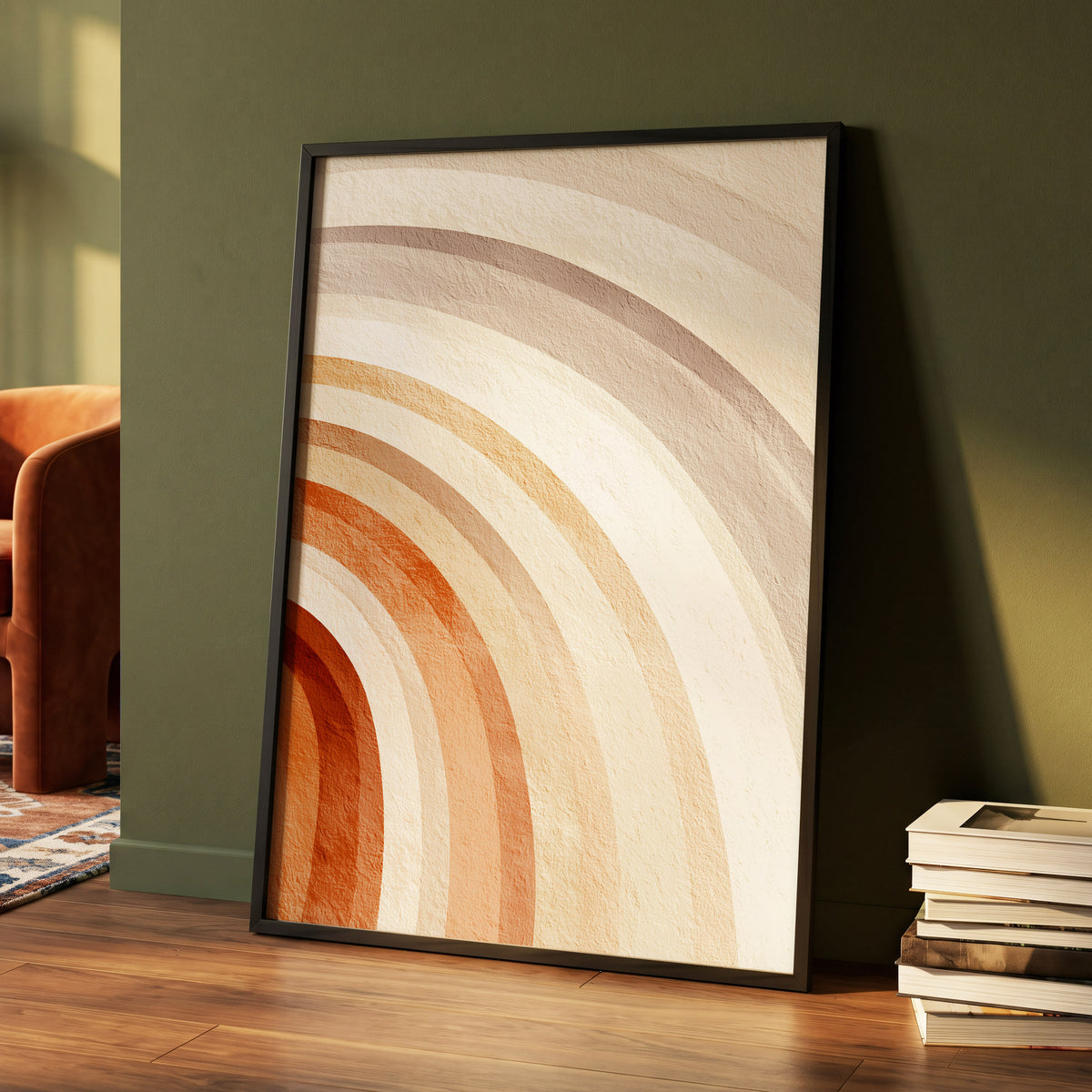 Terracotta Rainbow Arches: a framed abstract wall art print with concentric arches in cream, beige, tan, and rust orange, leaning against a dark olive green wall on a wood floor next to a stack of books.